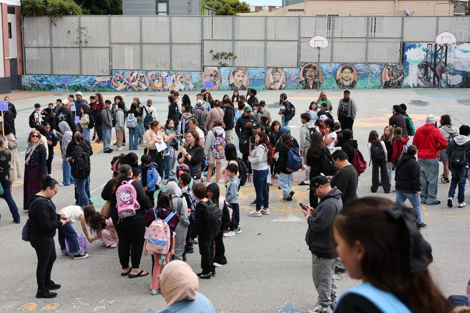 A large group of students and adults gather in a schoolyard with colorful murals on the wall and basketball hoops in the background.