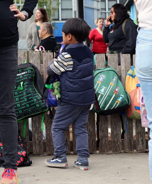 A young boy stands near a wooden fence with backpacks hanging on it, while adults and children are present in the background.