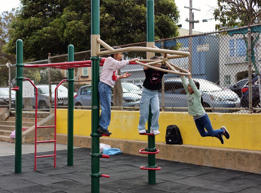 Three children play on a playground climbing structure; two are holding on to bars while one swings, with a yellow wall and parked cars in the background.