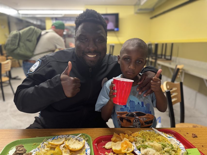 A man and a child sit at a table with plates of food; the man is smiling and giving a thumbs up, while the child holds a red cup and looks at the camera.