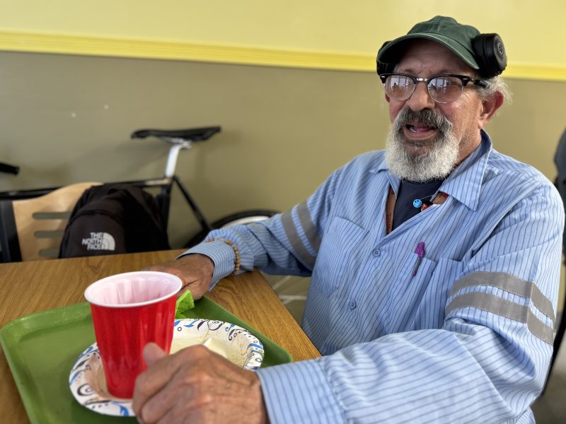 An older man with a gray beard and glasses sits at a table with a red cup and paper plate on a green tray; a bicycle is visible in the background.