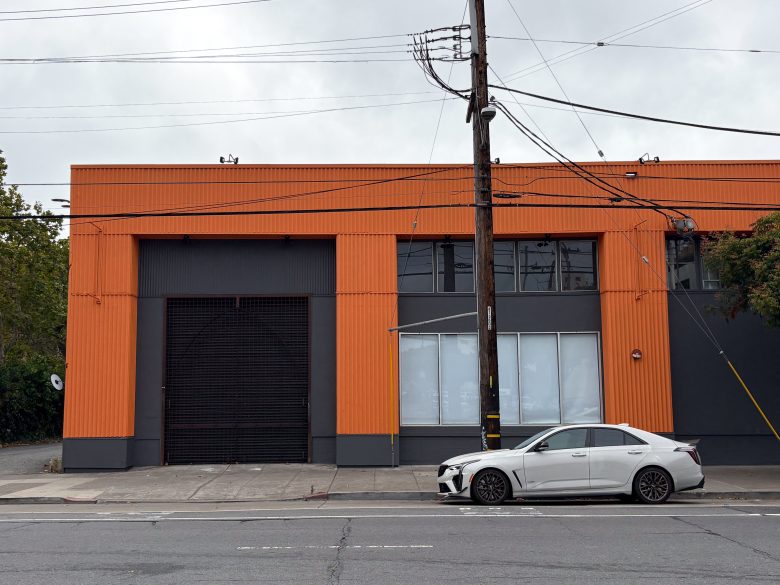 A white car is parked in front of an industrial building with orange and dark gray siding on a cloudy day.