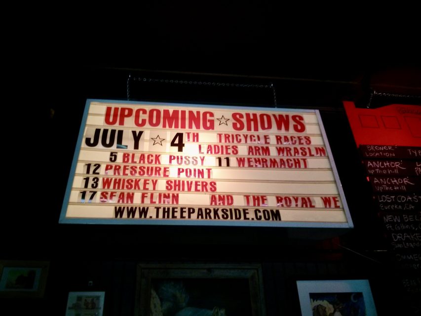 A marquee sign displays upcoming shows in July, including Tricycle Races, Ladies Arm Wrasslin, Black Pussy, Wehrmacht, Pressure Point, Whiskey Shivers, and Sean Flinn.