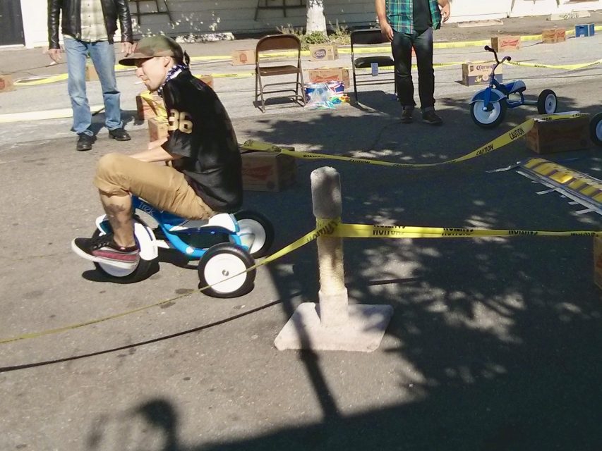 An adult man rides a blue tricycle in a cordoned-off area outdoors, with two other men standing nearby and folding chairs and caution tape visible.