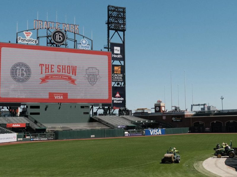 Grounds crew maintains the field at Oracle Park as a large scoreboard displays "THE SHOW" and various sponsor logos in an empty baseball stadium.