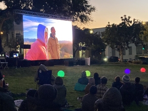 People sit on grass watching a movie projected on a large outdoor screen at dusk, with glowing colored lights scattered around the lawn.