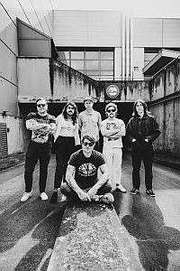 Six people pose for a black-and-white group photo outside an industrial building; five stand while one sits on a concrete path in front of them.