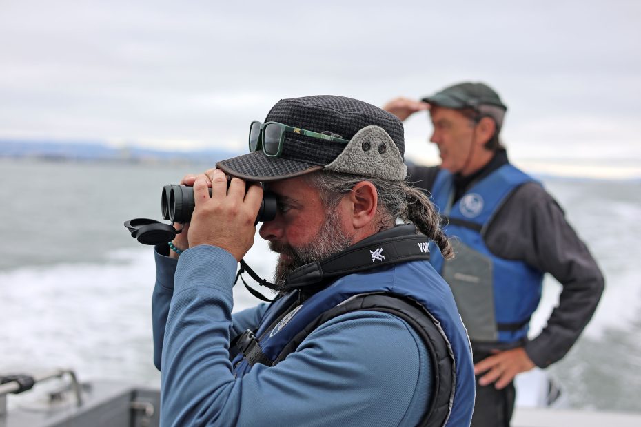 Two men on a boat, one looking through binoculars and the other shading his eyes with his hand, both wearing life jackets and hats.