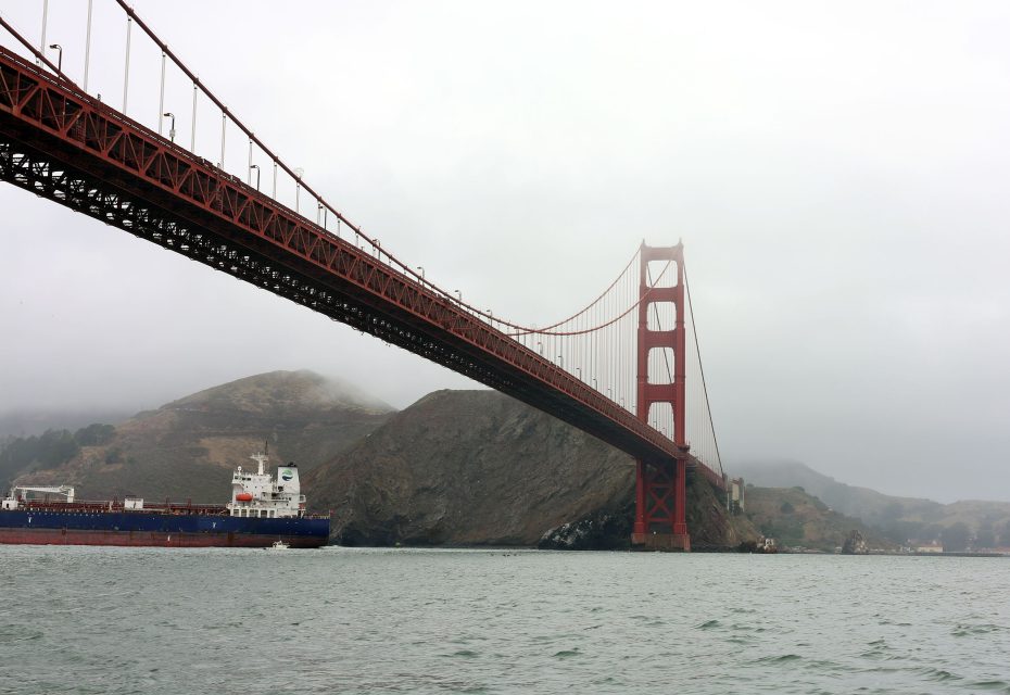 A large cargo ship passes under the Golden Gate Bridge on a cloudy day, with hills visible in the background.
