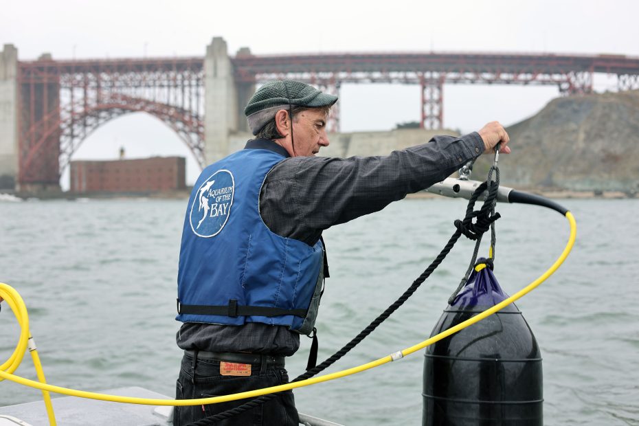 A man in a blue life vest handles equipment on a boat in the water, with a large red bridge and buildings visible in the background.