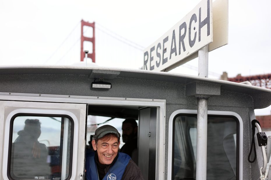 A man smiles while standing in the doorway of a research boat on the water, with a large "RESEARCH" sign and the Golden Gate Bridge visible in the background.