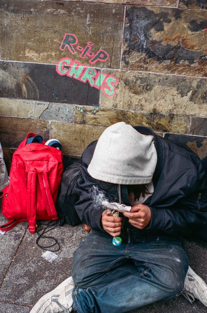 A person in a gray hoodie sits on the ground against a wall, smoking from a pipe. A red backpack and shoes are beside them. "RIP CHRIS" is written on the wall in colorful letters.