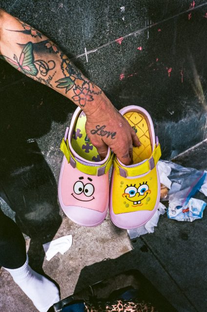 A tattooed hand holds a pair of cartoon-themed clogs featuring Patrick Star and SpongeBob SquarePants, with litter on the ground nearby.
