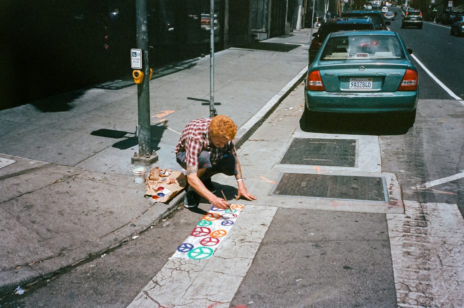 A person paints colorful peace symbols on a crosswalk at a city street corner beside a green car and a traffic signal pole.