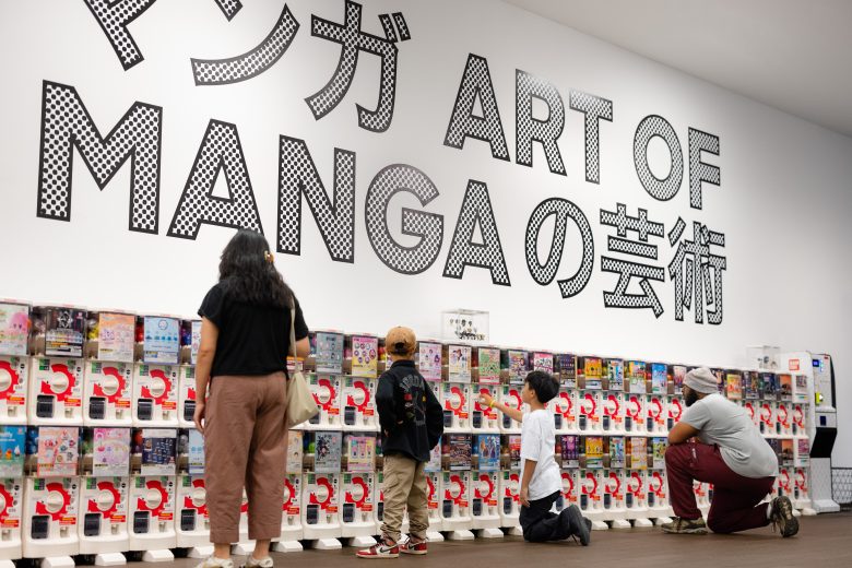 People look at and interact with rows of capsule toy machines in front of a wall displaying large text reading "ART OF MANGA" in English and Japanese.