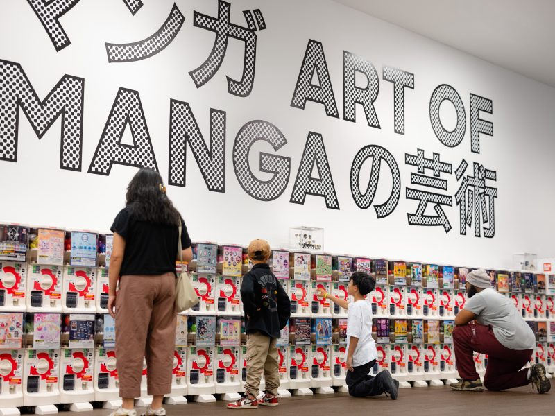 People look at and interact with rows of capsule toy machines in front of a wall displaying large text reading "ART OF MANGA" in English and Japanese.