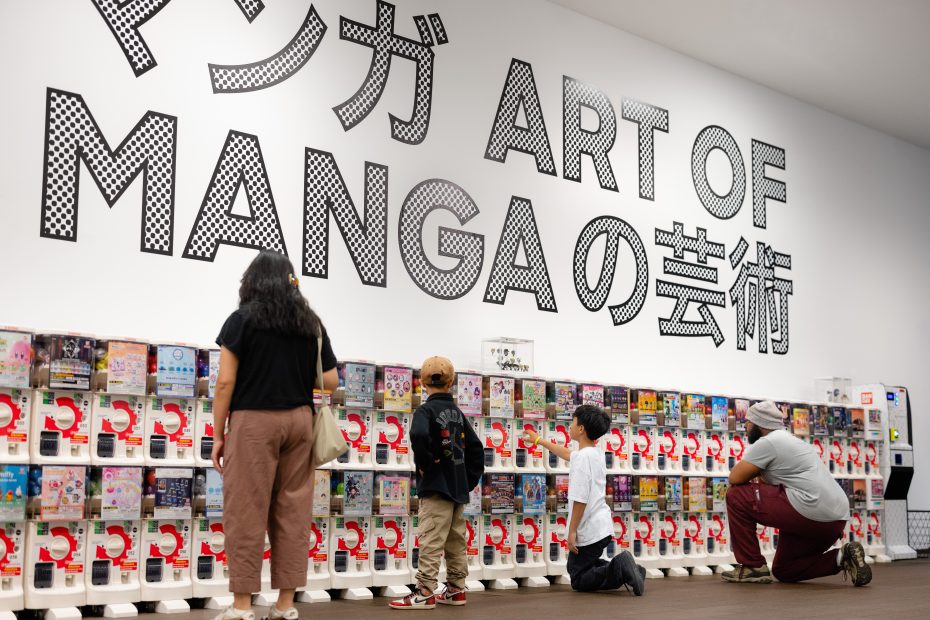 People look at and interact with rows of capsule toy machines in front of a wall displaying large text reading "ART OF MANGA" in English and Japanese.
