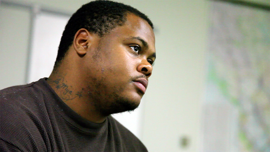 A man in a dark shirt with a neck tattoo looks to the side in a brightly lit room with a blurred background.