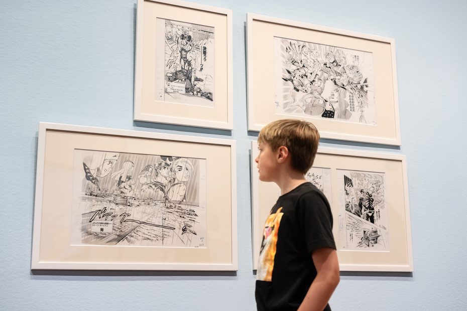 A boy stands in front of a wall displaying four framed black and white comic-style drawings.