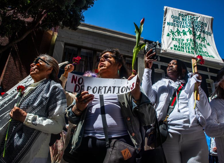 A group of people hold flowers and protest signs, including one that says “NO DEPORTATIONS” and another advocating to keep families together.