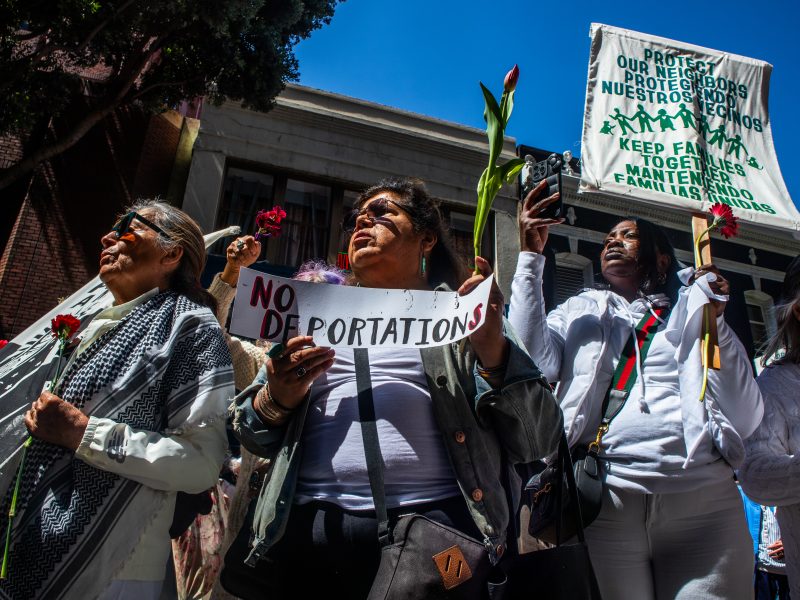 A group of people hold flowers and protest signs, including one that says “NO DEPORTATIONS” and another advocating to keep families together.