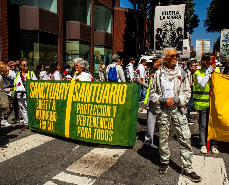 A group of people stand at a crosswalk holding a large banner reading “Sanctuary: Safety & Belonging for All / Santuario: Protección y Pertenencia para Todos” during a protest.
