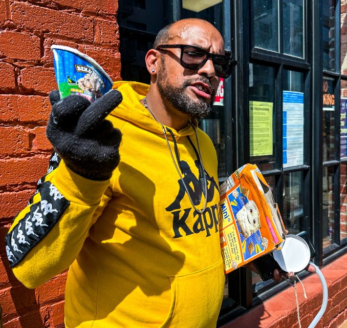 A man wearing sunglasses and yellow hoodie holding food and a bag of Frosted Mini Wheats.
