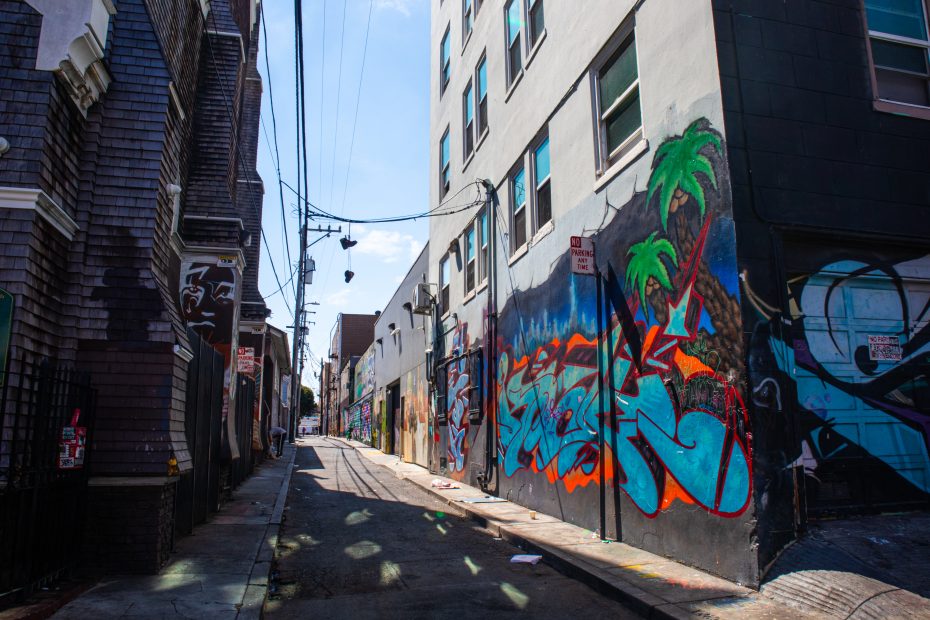 A narrow urban alleyway with colorful graffiti, including palm trees, covers the wall of a building on the right; other buildings and power lines line the alley.