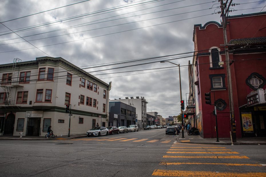 A city street intersection with crosswalks, parked cars, traffic lights, and multi-story buildings under a cloudy sky.