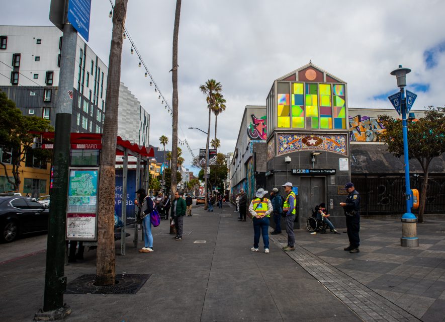 People wait near a bus stop on a city street with palm trees, colorful buildings, and overcast skies. Some are standing, while others are sitting or talking.