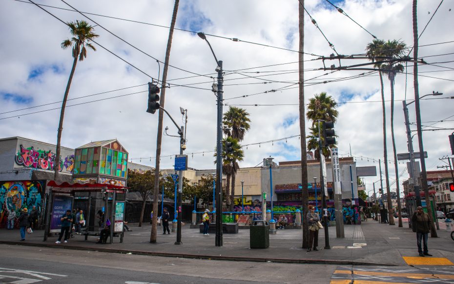 Urban street scene with palm trees, power lines, and buildings covered in colorful graffiti and murals; people are walking and waiting near a bus stop.