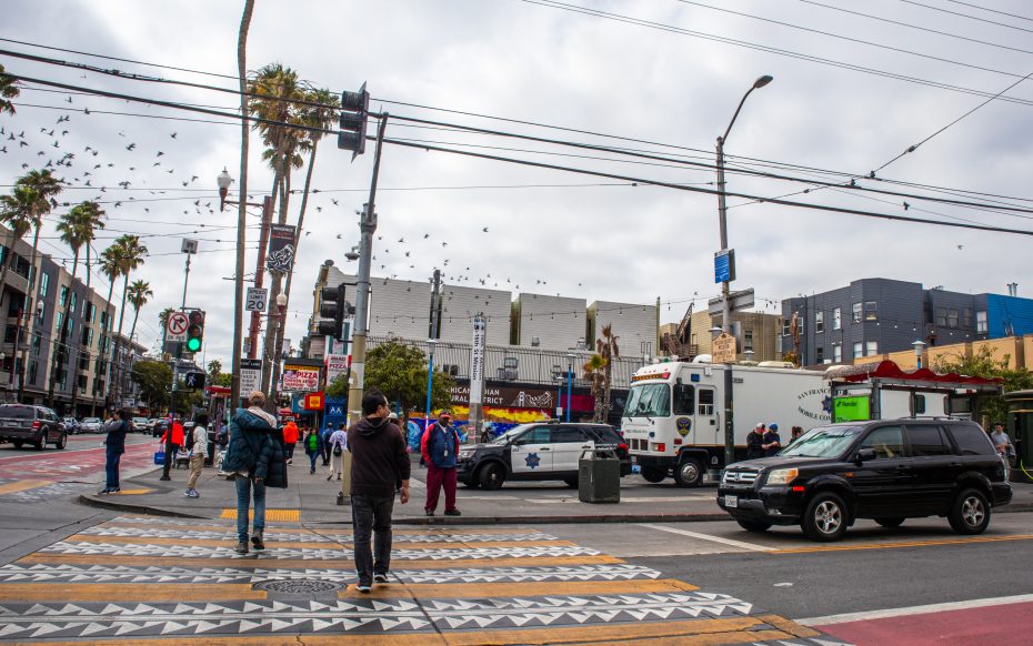 People cross a city street at a busy intersection with police and an armored truck present; buildings, palm trees, and overcast sky in the background.