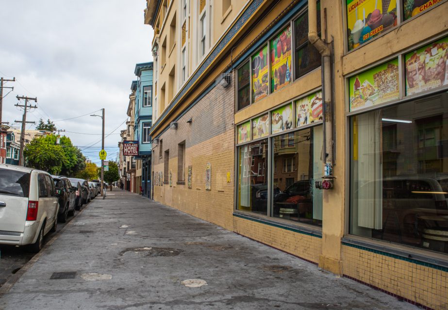 A city sidewalk with parked cars on the left and a building with large windows and colorful posters on the right under a cloudy sky.