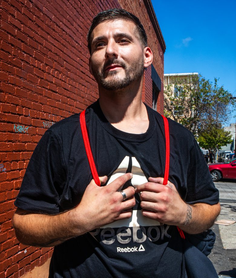 A man standing outside against a red brick wall, wearing a black Reebok t-shirt and holding red backpack straps, with buildings and a red car in the background.
