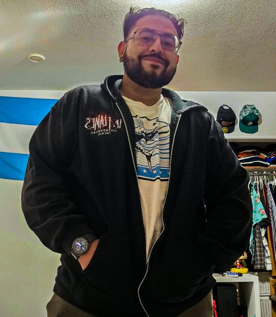 Man wearing glasses, a black zip-up jacket, and a printed white t-shirt stands with hands in pockets; a closet and a blue and white flag are visible in the background.