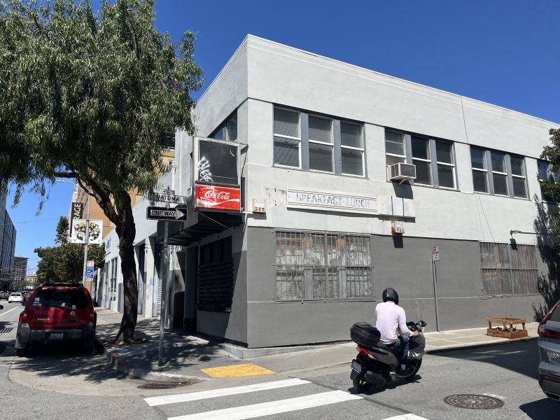 A person rides a scooter past a gray, two-story building with boarded windows on a sunny street corner.