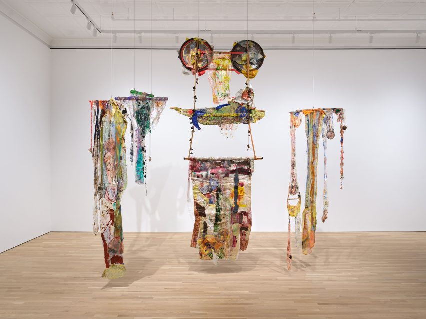 Mixed-media hanging sculpture with colorful, translucent fabric, plastic, and found objects suspended from the ceiling in a white-walled gallery with a wooden floor.