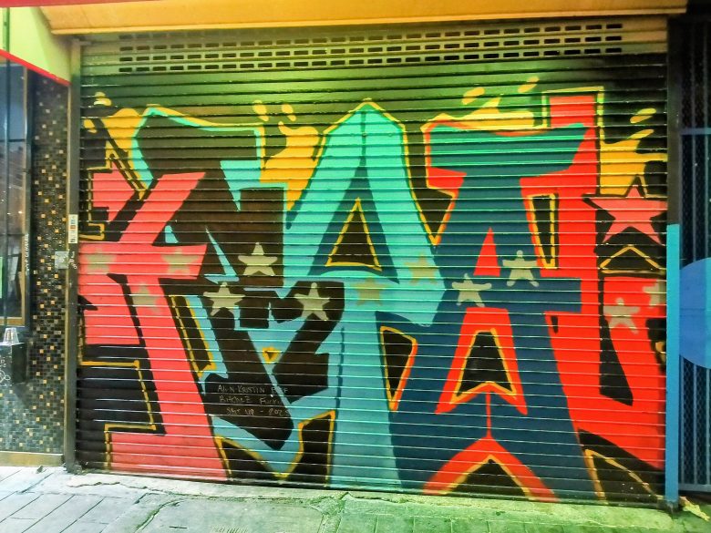 A metal shop shutter covered with colorful graffiti art featuring bold red, yellow, teal, and black shapes, with stars integrated into the design.