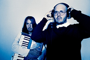 Two men pose in front of a light background; one holds a keyboard, the other wears headphones and glasses.