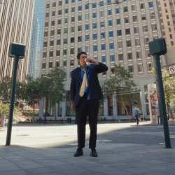 A man in a suit with a loose tie stands in an urban plaza, drinking from a cup, surrounded by tall office buildings.