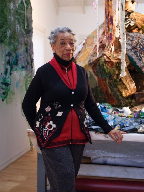 An older woman with short gray hair stands indoors, wearing a red top and black sweater, next to a table with colorful art materials and a large mixed-media sculpture.