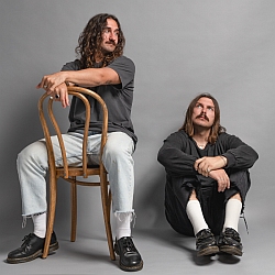 Two men with long hair and mustaches pose in a studio; one sits backward on a wooden chair, the other sits on the floor with arms on knees, both wearing casual clothes.