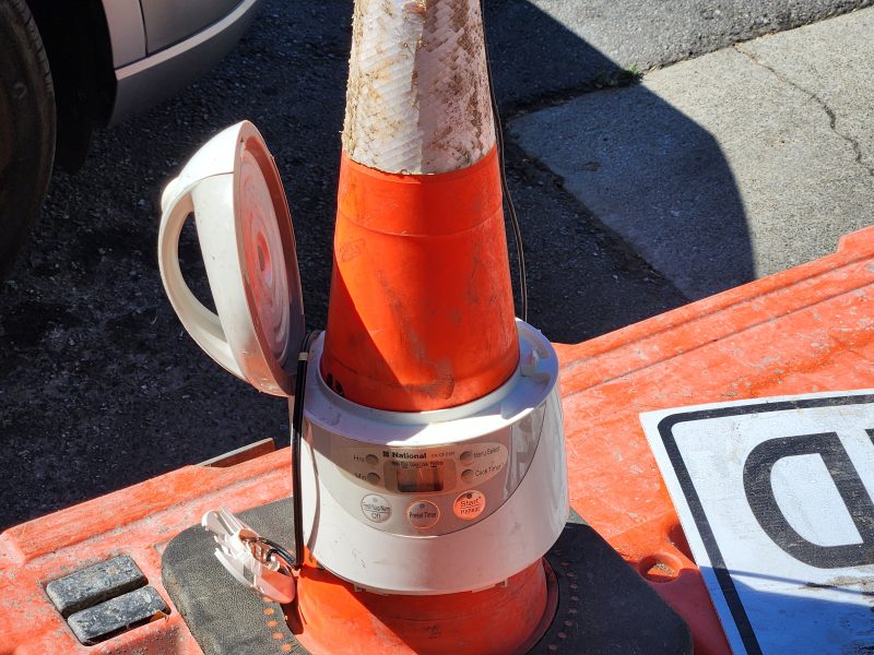 An electric rice cooker with its lid open is fitted around the base of an orange traffic cone on a plastic barrier near a street.