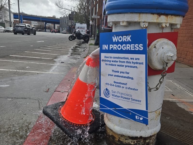 A blue fire hydrant leaking water at the base, with an orange traffic cone and a "Work in Progress" sign from San Francisco Water Power Sewer attached.