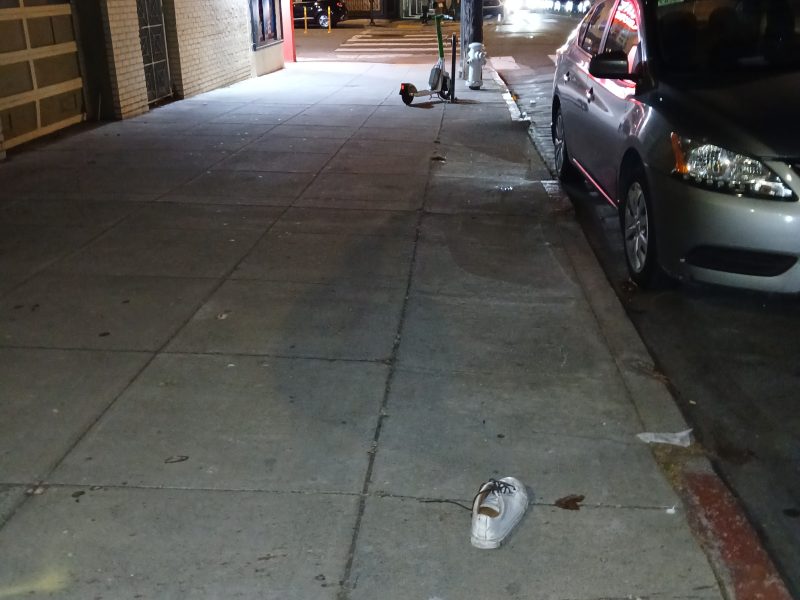 A pair of white sneakers is left abandoned on a city sidewalk at night, with a silver car parked nearby and a market storefront visible in the background.