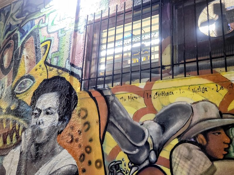 Colorful mural under a barred window, featuring a grayscale portrait of a person exhaling smoke and another person wearing a hat, with abstract and animal-like shapes in the background.