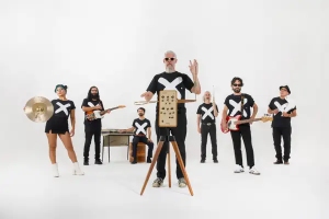 Seven people stand in a row on a white background, each wearing a black outfit with a white X. They hold various musical instruments, including a theremin, cymbal, guitar, and bass.