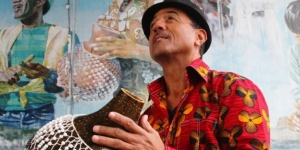 A man in a red patterned shirt and black hat holds a shekere instrument, standing in front of a colorful mural.