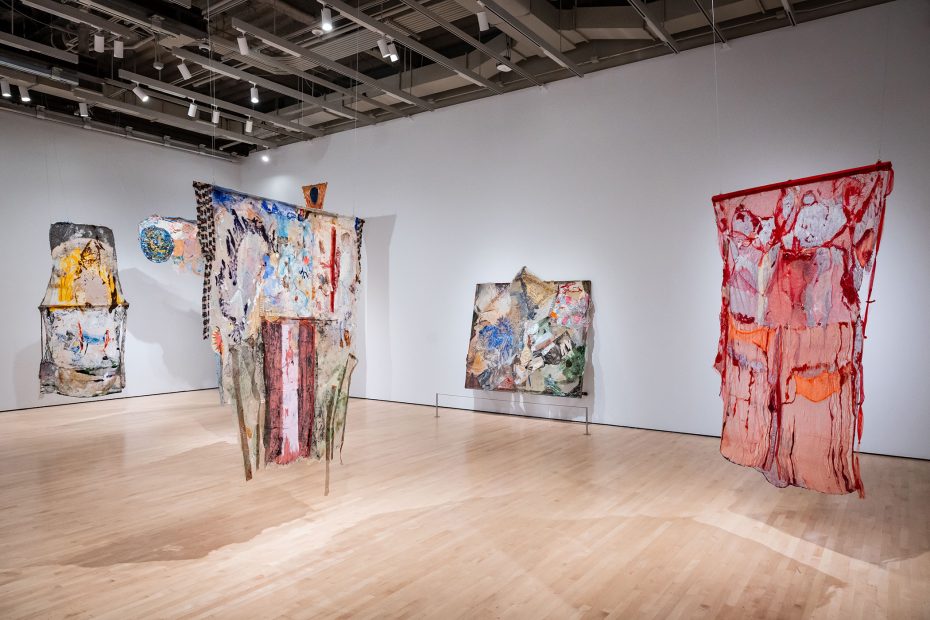 Contemporary art gallery with four large abstract mixed-media artworks displayed on white walls and wooden floor under ceiling lights.