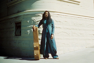 A person with long hair stands on a sidewalk, leaning on a guitar case, next to a light-colored stone building.